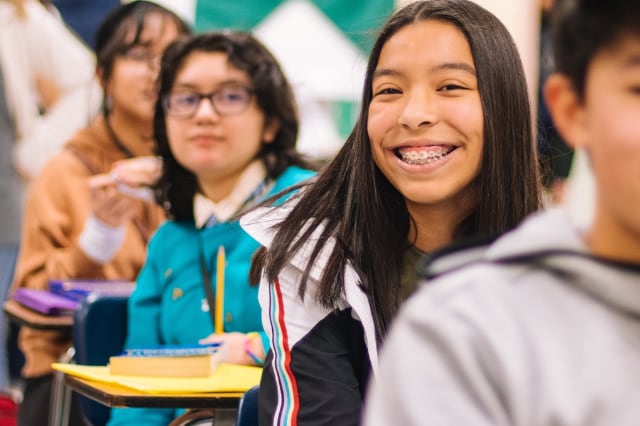 A happy female student smiles at the camera while sitting at her desk surrounded by other students in a classroom environment. 